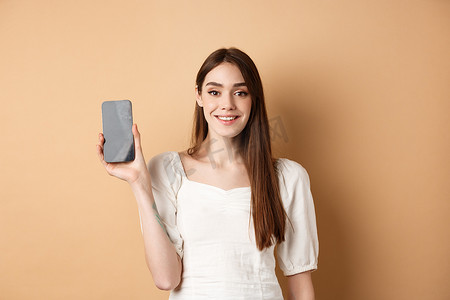 app展示摄影照片_快乐的女孩展示空的手机屏幕，微笑着，展示手机应用程序，站在米色背景上