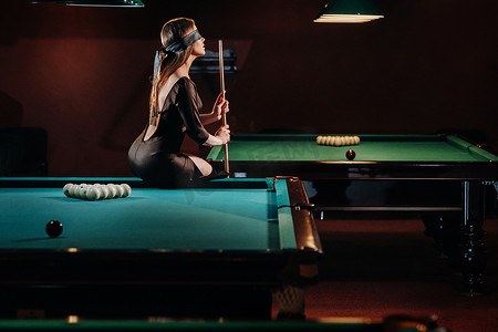 一个蒙着眼睛、手里拿着球杆的女孩坐在台球俱乐部的桌子上。俄罗斯台球