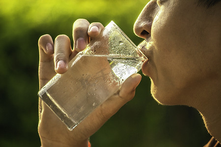 亚洲青少年男孩在模糊的自然背景下用透明玻璃杯喝凉水。