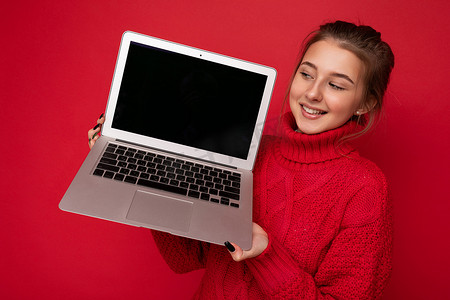照片中，美丽、满意、快乐的年轻女子拿着电脑笔记本电脑，看着显示器，穿着红色毛衣，隔离在红墙背景上