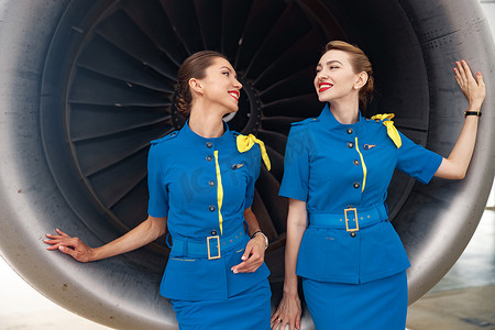 两位穿着时尚蓝色制服的漂亮空姐互相微笑，一起站在飞机发动机前