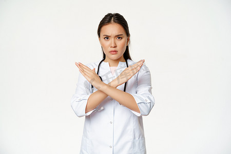 严肃的亚洲女医生禁止病人行为，表现出停止，交叉双臂手势，皱眉不悦，不赞成坏事，站在白色背景上