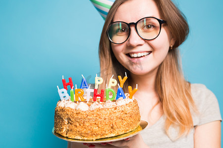 生日蛋糕海报摄影照片_戴着眼镜、问候纸帽、手里拿着一个生日快乐蛋糕、站在蓝色背景上的滑稽积极女孩的特写。