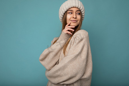 光荣背景摄影照片_一张漂亮快乐的年轻深色金发女子站在蓝色背景墙上的照片，她穿着米色温暖的毛衣，戴着冬季米色帽子，看着旁边，有一个想法