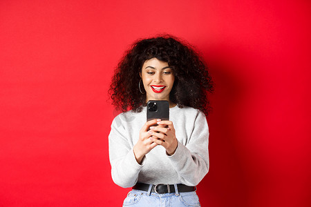 微笑的现代女孩在智能手机上拍照，看着屏幕并录制视频，站在红色背景下