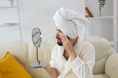 英俊的男人用棉垫清洁面部皮肤，看着镜子。