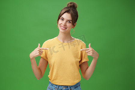 身穿黄色 T 恤、冷静、礼貌、友善、技术精湛的年轻女同事指着自己自信的倾斜头，对着镜头微笑，希望参加绿色背景的活动