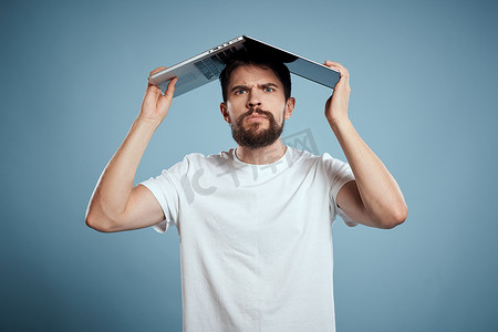 公众号台头摄影照片_情绪化的男人，头顶上有一台打开的笔记本电脑，蓝色背景，穿着白色 T 恤，裁剪视图