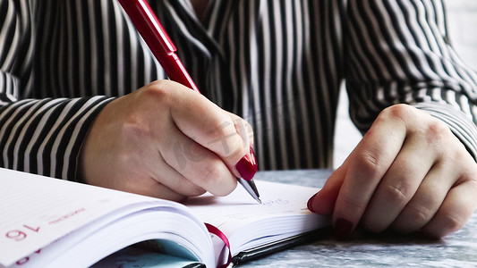 一个红指甲的女人的手的侧视图，拿着一支红笔，在一张灰色桌子上的空日记纸上写了一些东西。