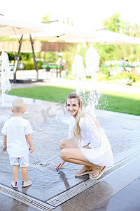小孩眼睛摄影照片_年轻快乐的母亲在喷泉附近和小孩玩耍。