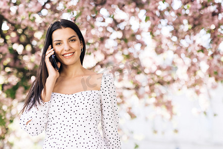 身穿白色夏装的年轻模特女孩在樱花树背景的公园里打电话聊天。