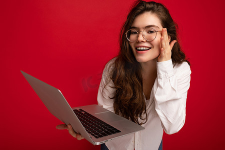 侧面照片中，美丽的微笑深色卷发年轻女子拿着电脑笔记本电脑，戴着眼镜白衬衫，看着红墙背景上孤立的上网本显示屏