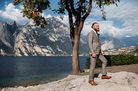 意大利加尔达湖附近阿尔卑斯山背景中，一位留着胡子的男子，身穿严格的灰色三件套西装，系着领带