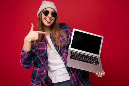 照片中，年轻女子拿着电脑笔记本电脑，戴着太阳镜帽子，穿着彩色衬衫，笑着笑着，看着相机露出舌头，指着红墙背景上隔离的上网本显示器