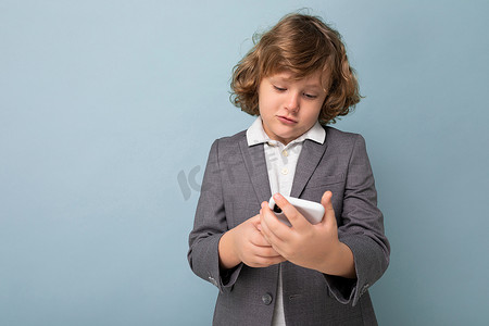 短卷发男孩摄影照片_照片中，英俊的小男孩，卷发，身穿灰色西装，手持并使用蓝色背景中隔离的手机，看着智能手机通信 vie 短信
