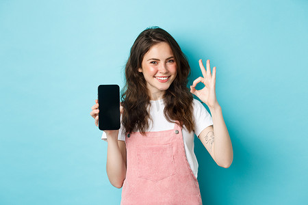 现代时尚女孩推荐网上商店或移动应用程序的肖像，显示带有空智能手机屏幕的良好标志，点头同意，满意地微笑，蓝色背景
