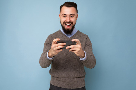 一张快乐、快乐、英俊、黑发、胡子拉碴的年轻男子的照片，他留着胡子，穿着日常的灰色毛衣和蓝色衬衫，隔离在背景墙上，拿着智能手机，通过手机玩游戏，看着手机屏幕显示，玩得开心