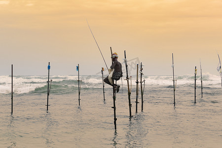 在日落的傍晚，高跷渔夫坐在他的竿子上，手里拿着一根木钓鱼竿。