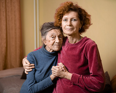 白种人老妇人温柔地拥抱她九十岁的老母亲，头发花白，脸上有深深的皱纹，在家里的沙发上，微笑着母女。