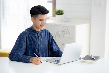 年轻的亚洲商人在笔记本上写作，规划工作，并在家里的办公桌上使用笔记本电脑，记录有关金融、男性学习、商业和沟通概念的笔记。