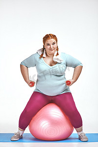 红发、胖乎乎的女人坐在球上，拿着哑铃