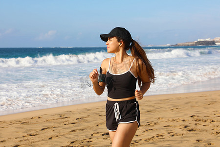 在夏日海滩上，戴黑帽的跑步女性在手机上听音乐，运动臂带配有触摸屏和耳机
