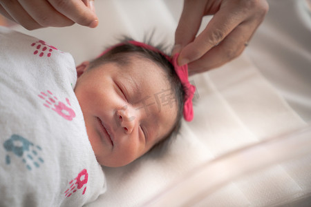 在医院里，一名刚出生的女婴侧躺在滚动摇篮车里，裹着襁褓毯，休息和睡觉，而她的母亲正在给她固定蝴蝶结或头带。