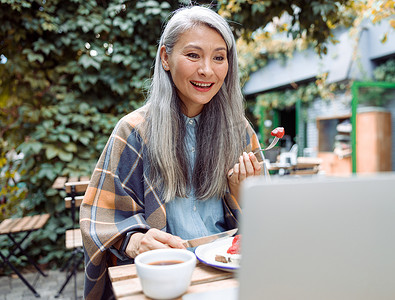 快乐的亚洲高级女士在户外餐桌上用笔记本电脑吃吐司和草莓，观看视频