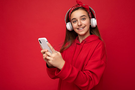美丽积极微笑的深色女青少年的特写肖像照片，身穿红色连帽衫，在红色背景下被隔离，手持并使用智能手机在线交流，戴着白色无线耳机，听着酷炫的音乐，看着相机
