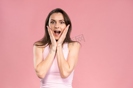 OMG 让穿着粉色 T 恤、双手放在脸上、脸上带着积极情绪的兴奋和喜悦的年轻漂亮女人感到惊讶。