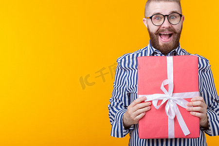 戴着眼镜、留着小胡子、快乐的年轻嬉皮士拥抱着一个黄色背景的红色礼盒，上面有复印空间。