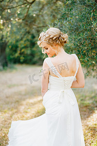一位温柔的金发新娘站在树林中间的一棵橄榄树旁，后视