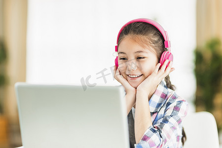 戴耳机的学生摄影照片_戴着耳机的快乐学生女孩。在线学习笔记本电脑听力课程。