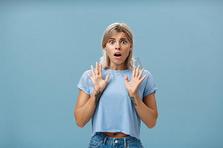 难过的女孩摄影照片_震惊和担心的漂亮女性，金发，纹身手臂，举起手掌，喘气，张开嘴，表达同情，为蓝色墙壁上的朋友感到难过