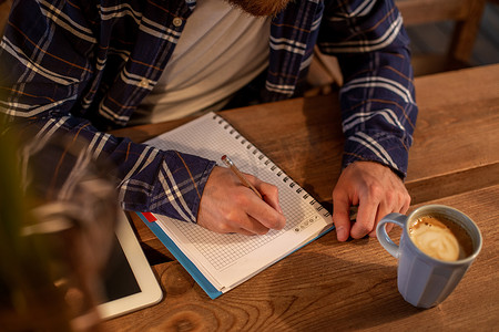 年轻的留着胡子的商人坐在咖啡馆里，坐在家里的桌子上，在笔记本上写字，附近有黑屏平板电脑。