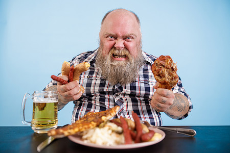愤怒的超重男子在餐桌上拿着鸡腿和香肠，上面放着油腻的食物