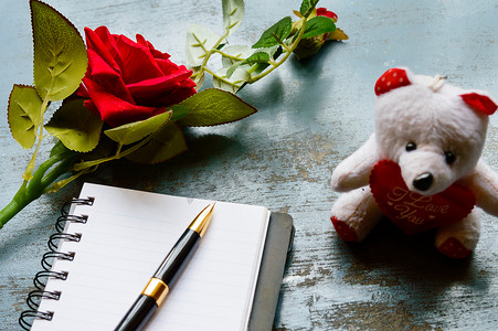 空白页笔记本、笔、一朵美丽的红玫瑰，放在泰迪熊附近的质朴金属地板背景上。