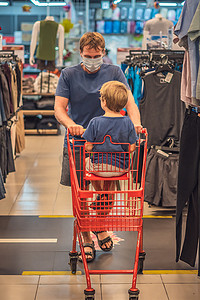 蓝色面罩摄影照片_严肃的父亲把小男孩卷进红色购物车，戴蓝色面罩，防止病毒感染，公共个人健康控制措施，大流行期间的家庭购物，购买新衣服