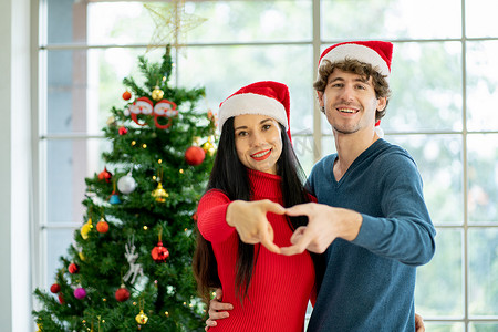 一对年轻男女用双手制作心的象征，他们幸福地呆在装饰着圣诞节的房间里。