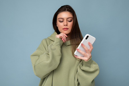 照片中，一张严肃、深思熟虑的美丽黑发年轻女子穿着时尚的绿色连帽衫，用手机在背景上隔离地写着信息，看着智能手机显示屏