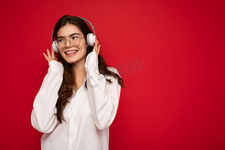 照片中，美丽快乐的黑发年轻女子身穿白衬衫，戴着光学眼镜，隔离在红色背景墙上，戴着白色无线蓝牙耳机，听着酷炫的音乐，欣赏着侧面