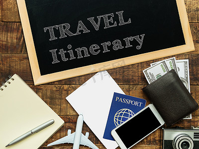 相机飞机摄影照片_黑板上用白色粉笔手写的旅行行程，在木质背景上装饰着飞机模型、护照、钱包、笔记本、相机和智能手机
