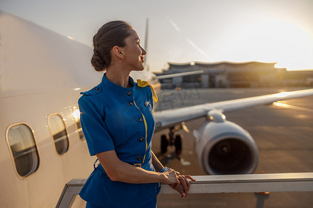 穿着蓝色制服的体贴的空姐看着别处，站在户外夕阳下。