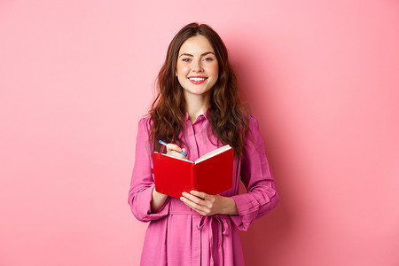 开朗的女孩微笑着写下笔记，拿着她的计划表、计划时间表或杂货清单，站在粉红色背景下的日记上