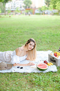 年轻漂亮的女孩读书，躺在格子上，靠近水果和帽子，背景是草。