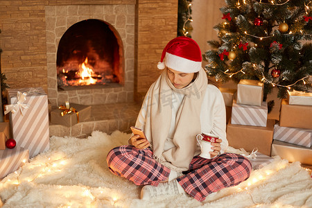 照片中，生病的女士坐在喜庆的客厅里，用手机拿着带有圣诞装饰品的饮料杯，给某人发短信，祝贺节日。