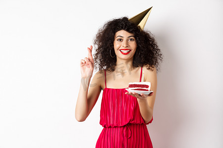 站着许愿的人摄影照片_兴奋的生日女孩庆祝、许愿、拿着蛋糕、交叉手指祝好运，在白色背景中快乐地站着