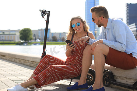 可爱的情侣在度假，享受驾驶电动滑板车的乐趣，开车休息一下，坐在河岸上
