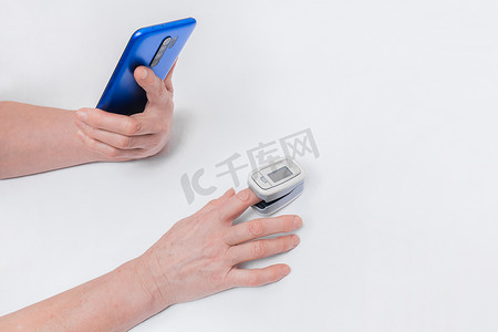 一位老年妇女的手握着手机或智能手机，用白色背景上的现代医疗设备脉搏血氧计测量血液中的饱和度和氧含量