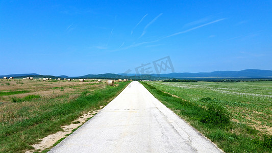 蓝天背景下夏末麦田干草卷之间的轻型乡村道路